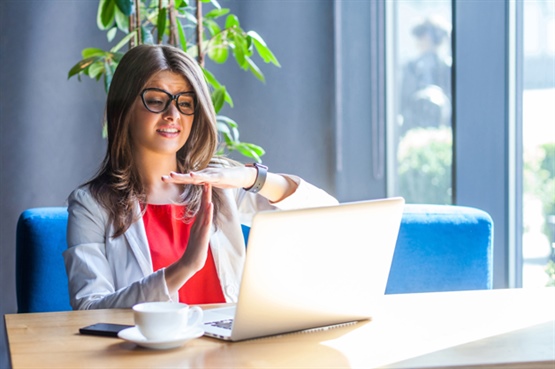 img-Zoom-fatigue3-555-370-p-L-97 woman wearing glasses sits at a desk, holding her hand up in a gesture, while using a laptop in a well-lit environment with plants in the background.