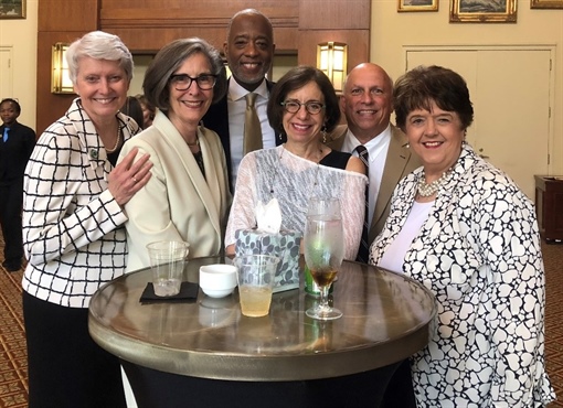 A group of six people standing together at a social event, smiling and posing for the camera with drinks on a table.